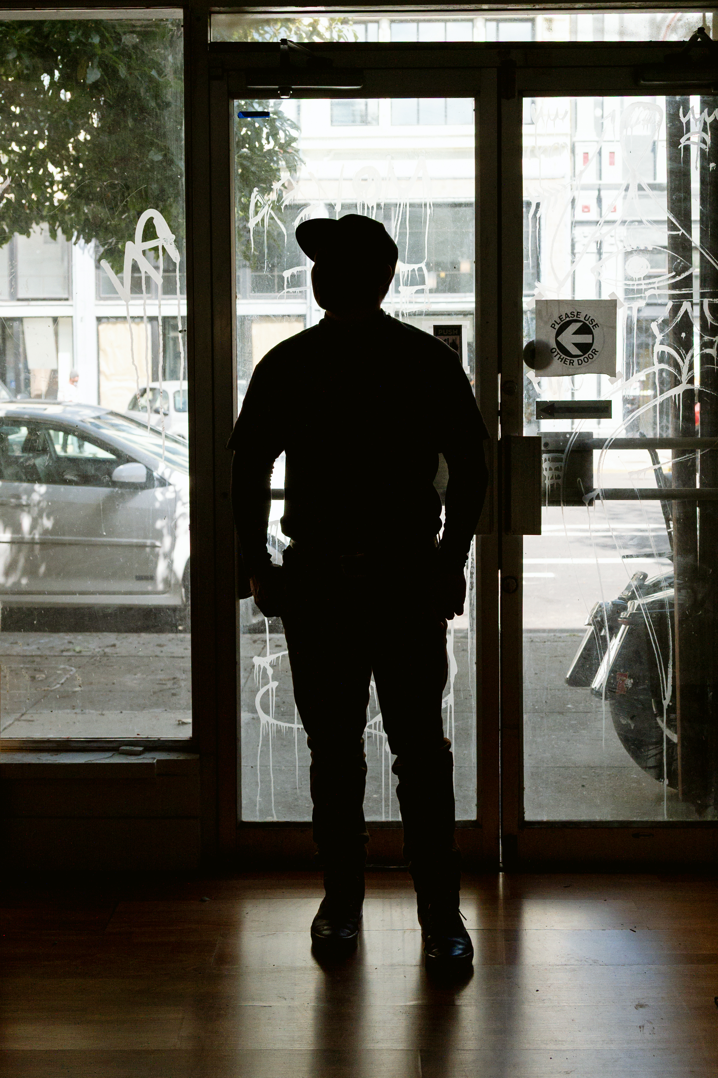 A person stands silhouetted in front of a glass door, facing outside onto a city street with cars and buildings visible through the glass.