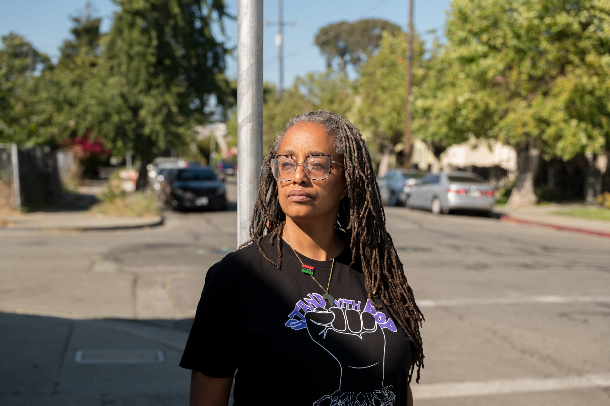 A person with long locs and glasses stands on a sunny street, wearing a black T-shirt that says “Stand with BOP” featuring a raised fist graphic.