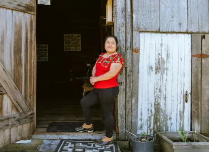 A smiling woman in a red embroidered blouse standing at the open doorway of a rustic wooden building.