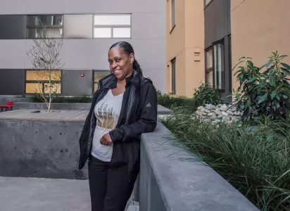 Sonja Sawyer wearing a jacket stands in a courtyard between apartment buildings, leaning against a concrete ledge surrounded by greenery.