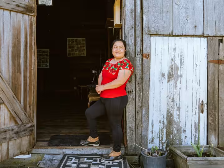 A smiling woman in a red embroidered blouse standing at the open doorway of a rustic wooden building.