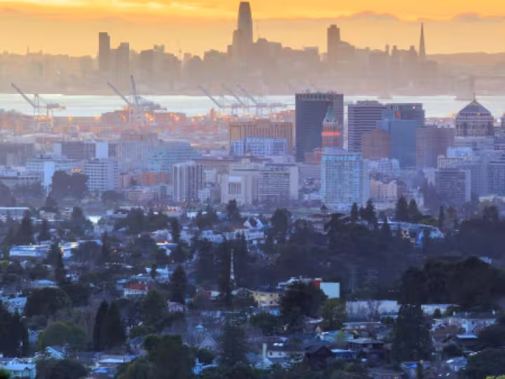 A view of the San Francisco Bay Area at sunset, showing urban buildings, cranes along the waterfront, and residential neighborhoods in the foreground.