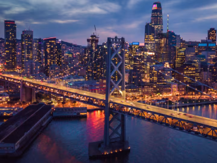 A nighttime aerial view of downtown San Francisco with the Bay Bridge lit up over the water.