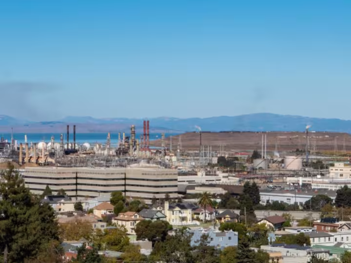 A view of a residential neighborhood in the foreground with an industrial refinery complex behind it, set against distant hills and a clear blue sky.