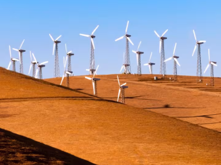 A row of wind turbines stands on rolling golden hills beneath a clear blue sky.