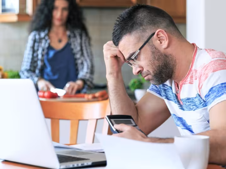 man looking at laptop and calculator while wife chops tomatoes