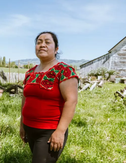 A woman wearing a red blouse with green and floral embroidery stands in a grassy field in front of a weathered white barn under a blue sky.