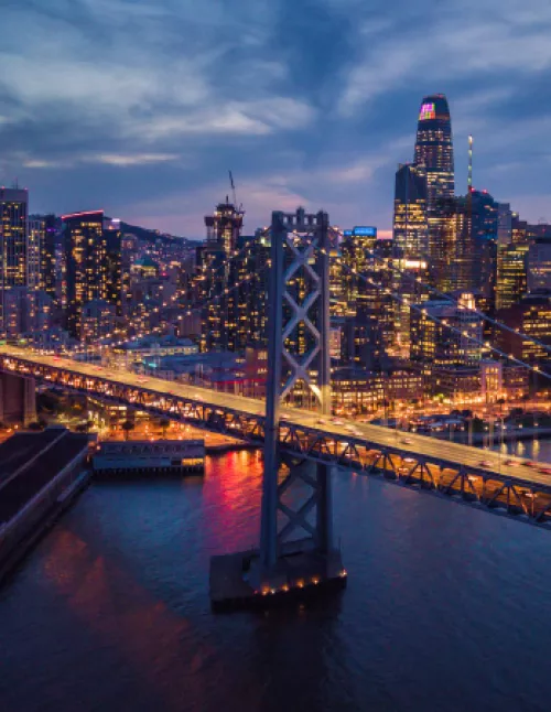 A nighttime aerial view of downtown San Francisco with the Bay Bridge lit up over the water.