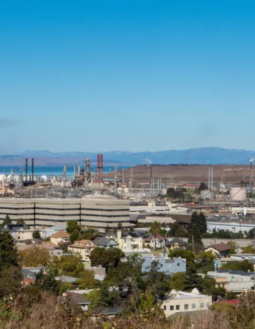 A view of a residential neighborhood in the foreground with an industrial refinery complex behind it, set against distant hills and a clear blue sky.