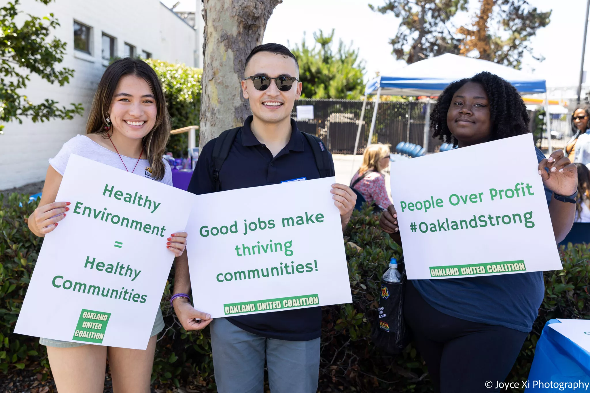 Three people smile while holding signs with community-focused messages such as “Healthy Environment = Healthy Communities,” “Good jobs make thriving communities!,” and “People Over Profit #OaklandStrong” at an outdoor event.